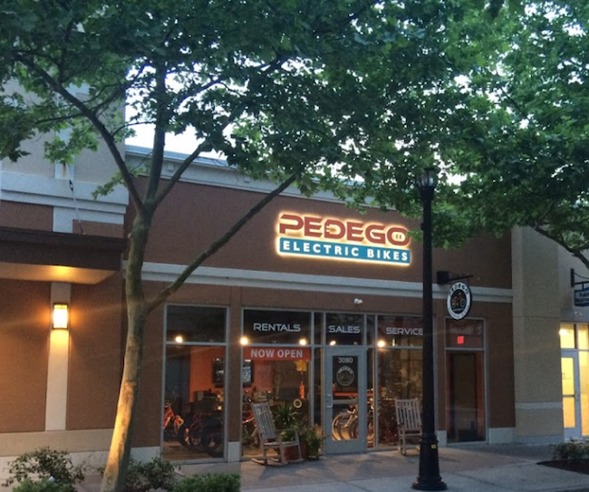 Front of store showing Pedego in Red lettering, Electric Bike in Blue & White letters, With Rentals, Sales Service on window in white lettering as well as showing the store with bikes inside it.