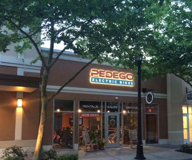 Front of store showing Pedego in Red lettering, Electric Bike in Blue & White letters, With Rentals, Sales Service on window in white lettering as well as showing the store with bikes inside it. 