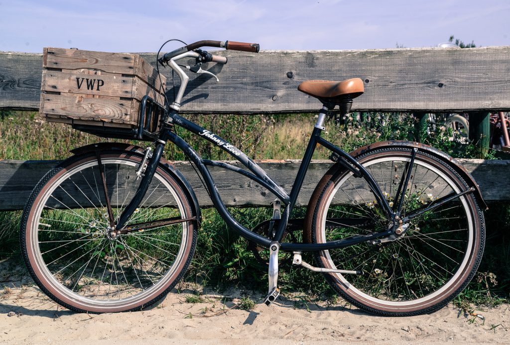 Black bicycle with tan seat and handlebars, wooden basket on front of handlebars leaning up against a wooden fence in sand. 